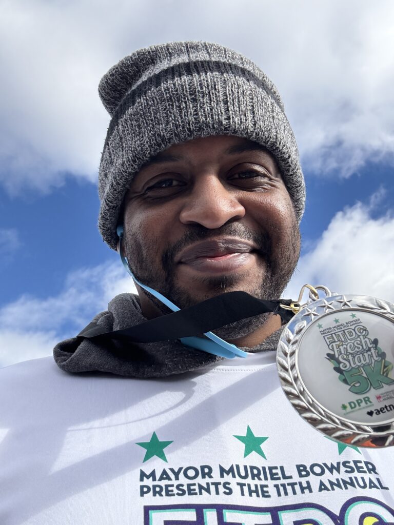 Zuri Berry, wearing a beanie and a white shirt, holds a medal up that reads "Fresh Fit 5K." Exercise is a big part of my 2025 resolutions.